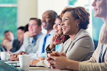 presentation Mid adult African American businesswoman is smiling while attending a job seminar or job training business conference. She s taking notes while listening to a speaker. She is dressed in smart business casual clothing and is sitting next to diverse group of professional coworkers.