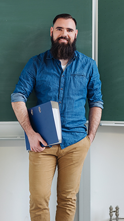 externships01 Relaxed confident male teacher in a classroom standing holding a binder in front of the chalkboard smiling at the camera