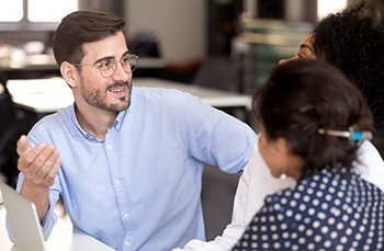 Team leader positive millennial man communicating with diverse female colleagues in office during meeting. Multiracial coworkers sitting together at desk talking discuss about new project share ideas