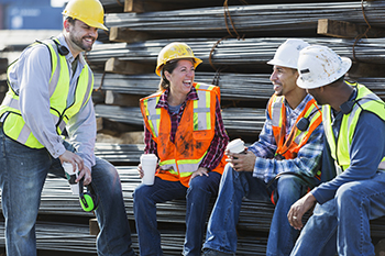 teachertour04 A multi-ethnic group of workers, a woman and three men, wearing safety vests and hardhats taking a coffee break. They are sitting on a stack of steel rebars (construction material), drinking coffee and talking.