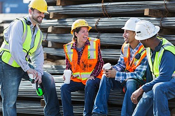 A multi-ethnic group of workers, a woman and three men, wearing safety vests and hardhats taking a coffee break. They are sitting on a stack of steel rebars (construction material), drinking coffee and talking.