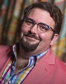 Head and shoulders shot of Corey Martin, a man wearing a dark pink suit jacket, a multicolored checked dress shirt and glasses.