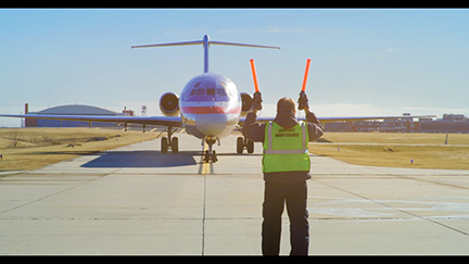 Man wearing a yellow safety vest stands in front of a plane to direct the pilot where to go.