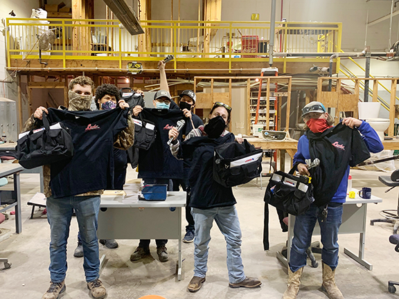 Mid-Del Technology Center students hold up black golf shirts, wrenches and black tool bags they received from Locke Supply. They are standing in a workroom.