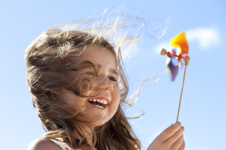 Little happy girl laughing and holding a pinwheel in front of a bright sky. The wind is blowing her hair illuminated by the sunlight. Conceptual image about environment and happiness
