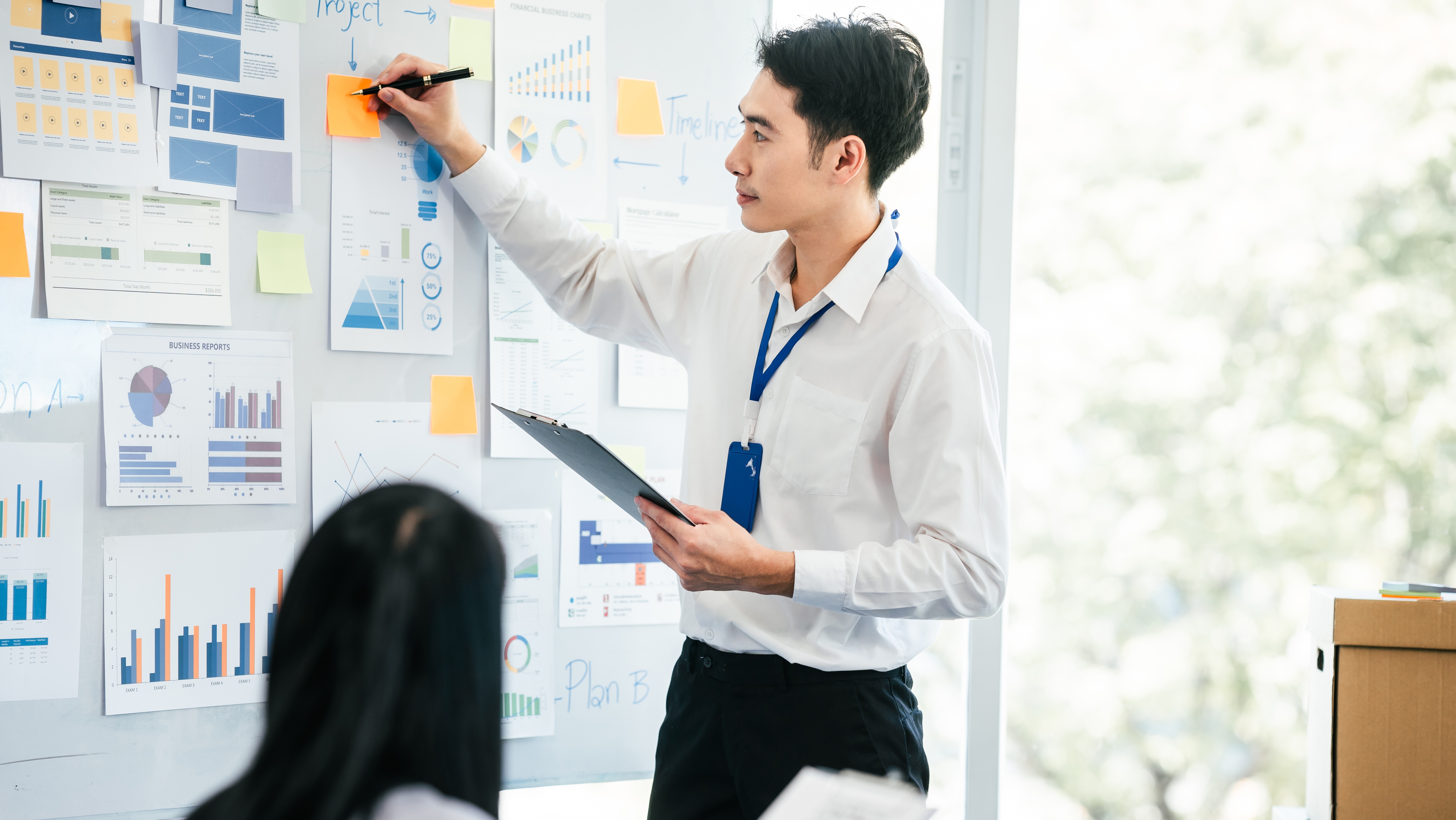 mature Asian business manager is leading a meeting, engaging with trainees in a bright room with data charts, indicating a monthly review or training session.