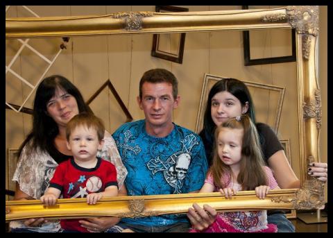 A wife and husband with three little girls hold a large empty frame around their family