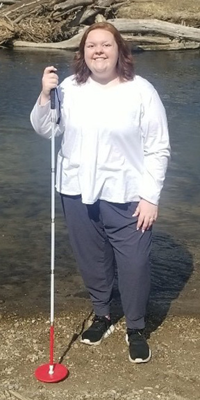 A woman with a white cane poses for the camera in front of stream.
