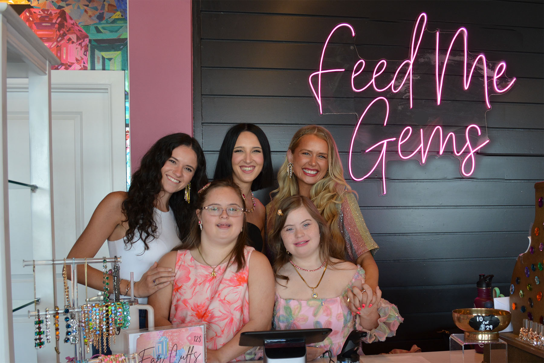 Five smiling women stand behind a store counter with words Feed me Gems on wall behind them. 