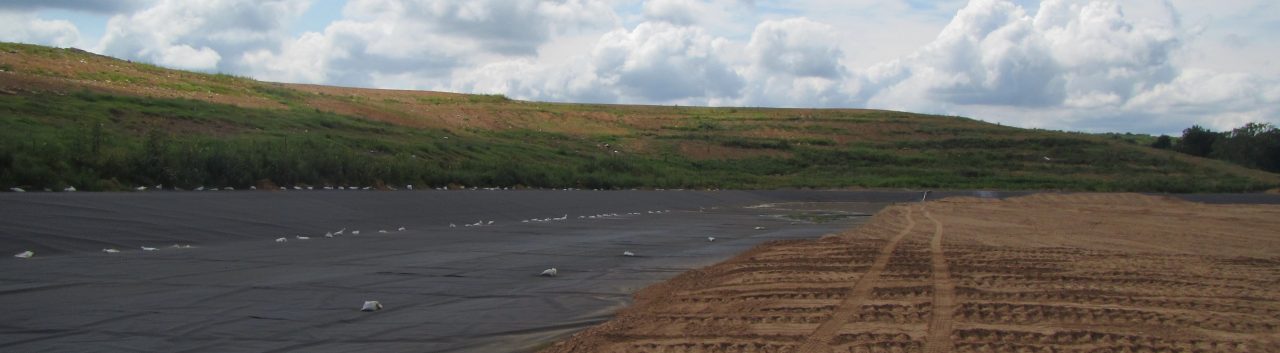 An image showing a cloudy day with green and brown pasture, a paved road, and an area with dirt and no grass.