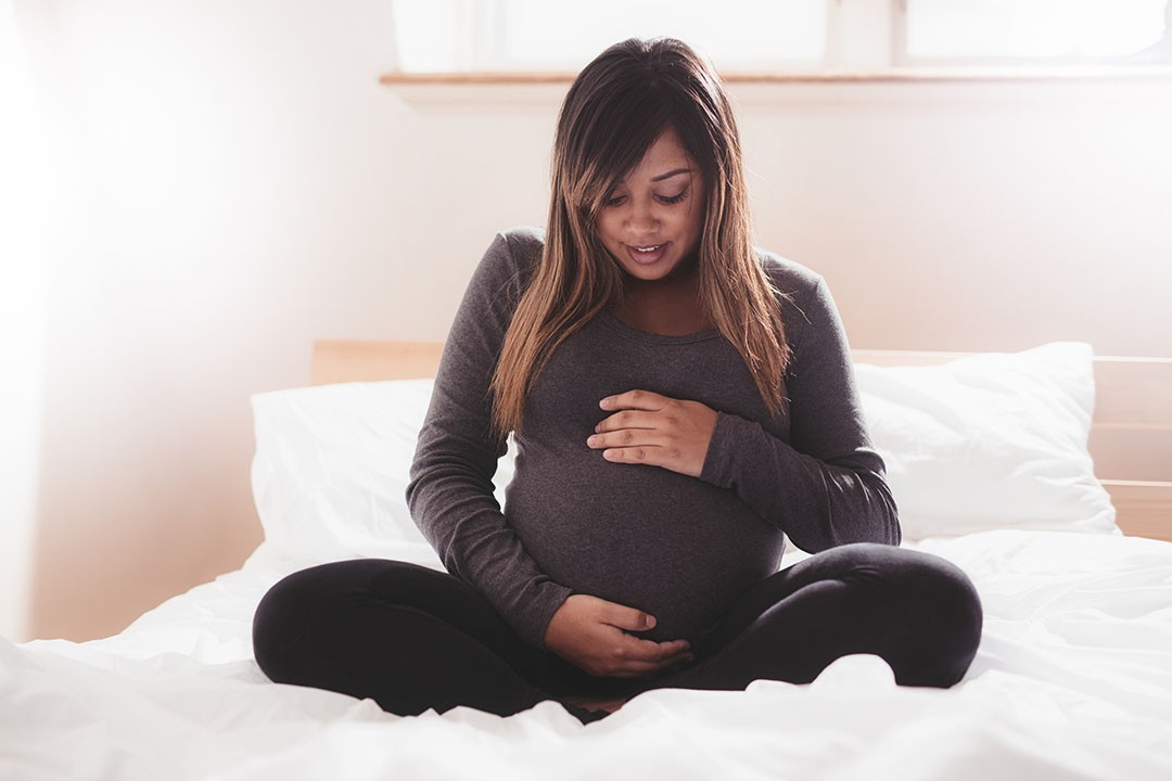 Beautiful pregnant woman sitting on bed at home. Young african pregnant woman relaxing in bedroom with her hands on her belly.