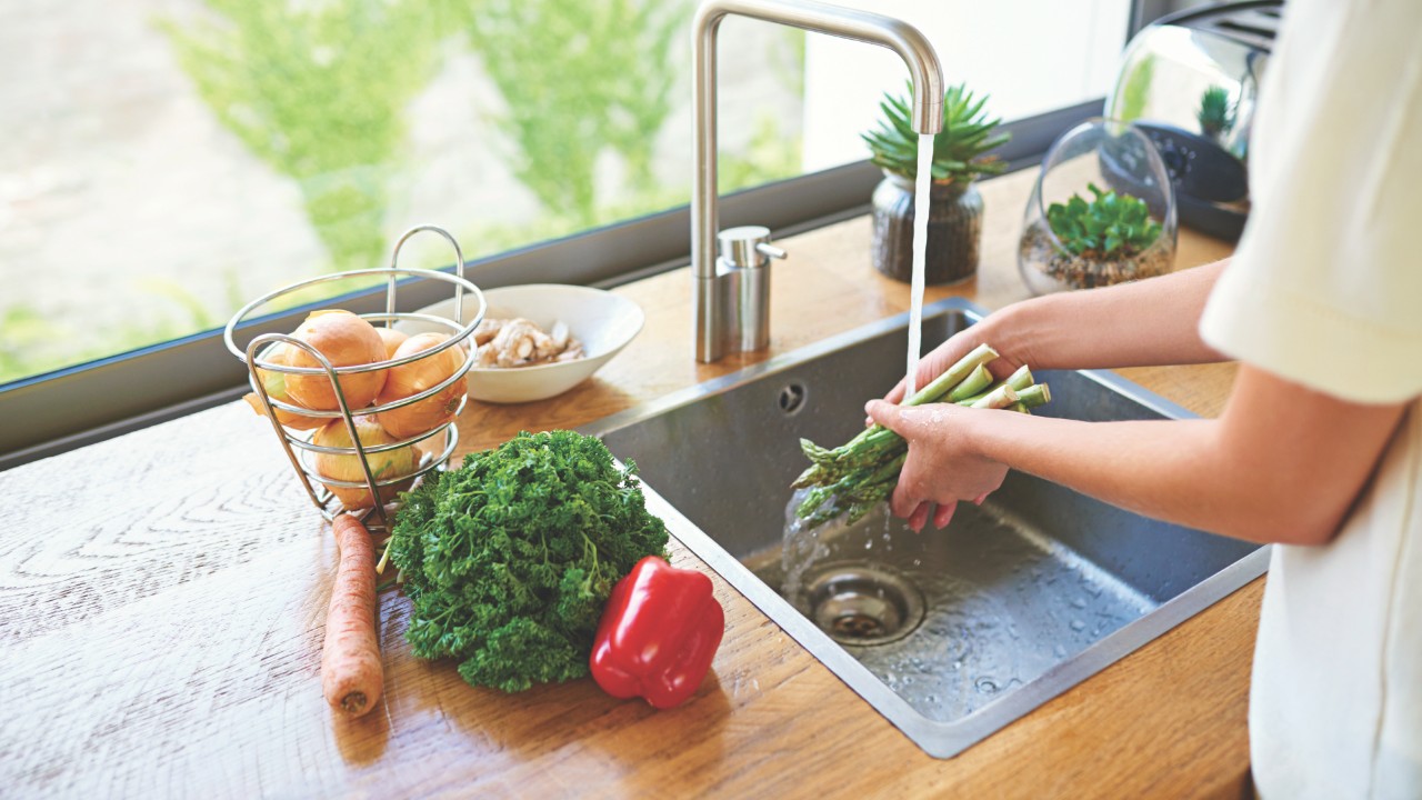 Person rinsing fresh asparagus under running water in a kitchen sink with vegetables on the counter.