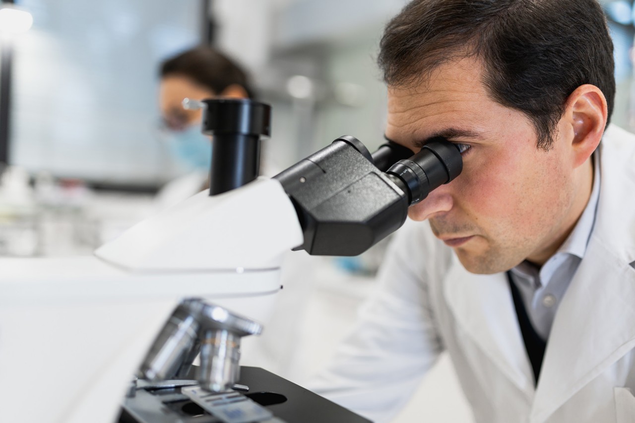 Concentrated male scientist using microscope and analyzing chemical samples while female colleague working on laptop in modern laboratory