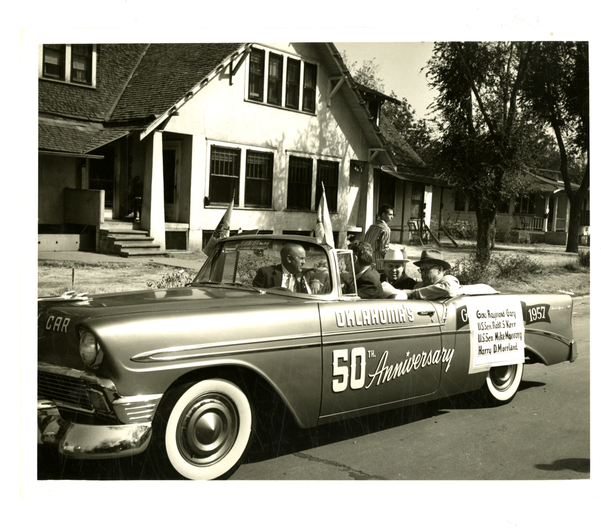 "Oklahoma's 50th Anniversary" car A black and white photo shows a vintage convertible car on a street, likely participating in a parade. The car has "Oklahoma's 50th Anniversary" painted on its side, along with "CAR" on the front fender and "1957" on the rear. Several men are visible in the car, possibly dignitaries, as a sign on the passenger side lists names: "Gov. Raymond Gary," "U.S. Sen. Robert S. Kerr," "U.S. Sen. Mike Monroney," and "Harry D. McParland." The car appears to be a Chevrolet, possibly from the mid-1950s, with prominent whitewall tires. In the background are houses and trees, suggesting a residential street.