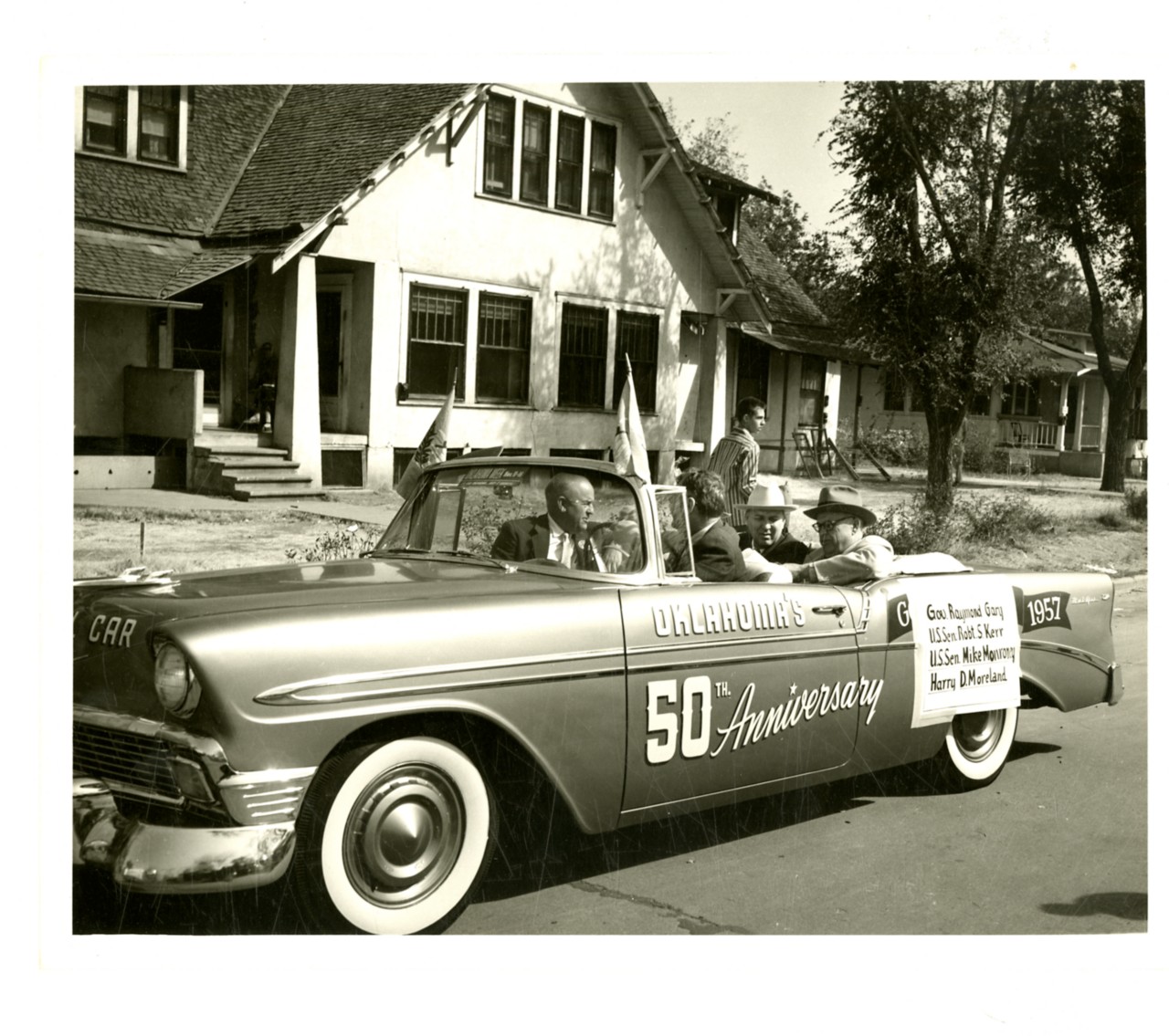 A black and white photo shows a vintage convertible car on a street, likely participating in a parade. The car has "Oklahoma's 50th Anniversary" painted on its side, along with "CAR" on the front fender and "1957" on the rear. Several men are visible in the car, possibly dignitaries, as a sign on the passenger side lists names: "Gov. Raymond Gary," "U.S. Sen. Robert S. Kerr," "U.S. Sen. Mike Monroney," and "Harry D. McParland." The car appears to be a Chevrolet, possibly from the mid-1950s, with prominent whitewall tires. In the background are houses and trees, suggesting a residential street.