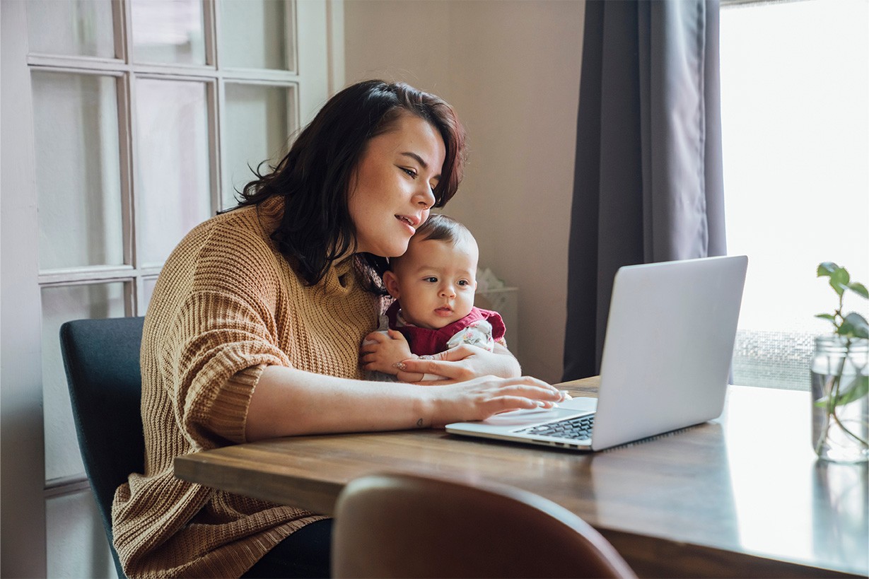 A mother holds a baby while using a laptop. 