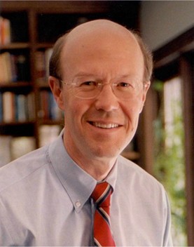 Head shot of Bob Anthony wearing glasses, blue shirt and red tie, smiling in front of bookcase