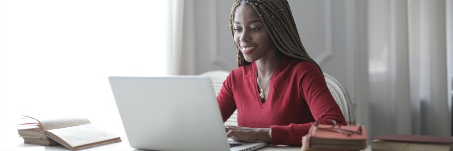 A woman with dark skin wearing her hair in braids and a red shirt smiles while typing on a white laptop