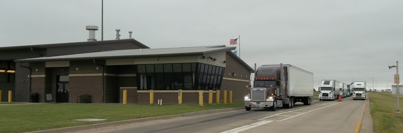 A semi-truck with a maroon tractor pulls up to a building that is the Port of Entry for the State of Oklahoma