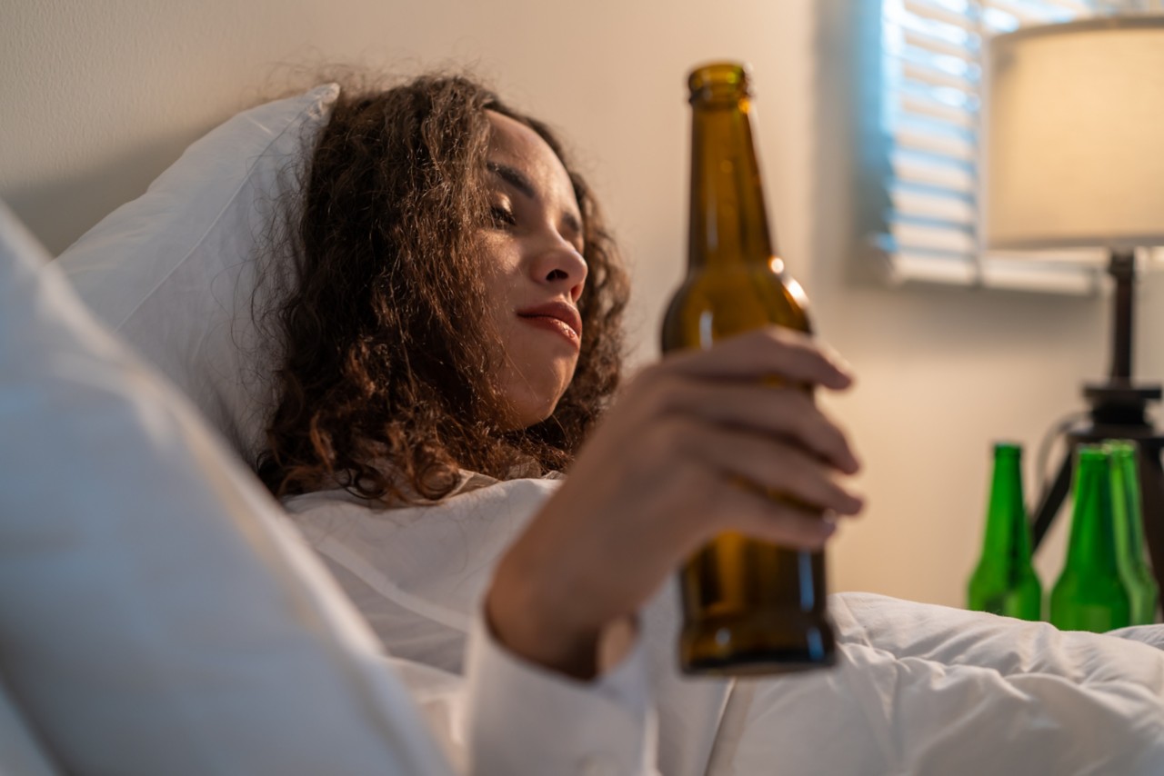 Drunk Latino woman hold beer bottle, and crying feel hangover on bed. Young beautiful depression female lying and crying in tears in bedroom, feel heart broken and celebrate life with alcohol at night