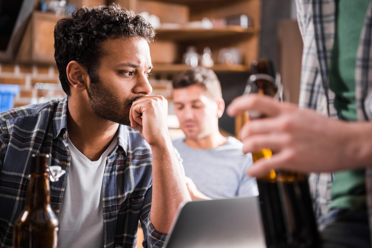 Serious young man using laptop while young people having fun with beer