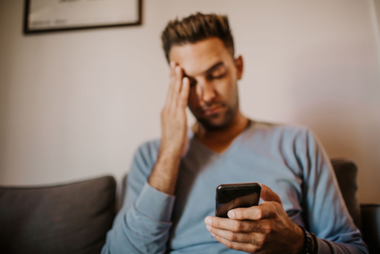 Young man sitting at home on sofa and using mobile phone looking distressed