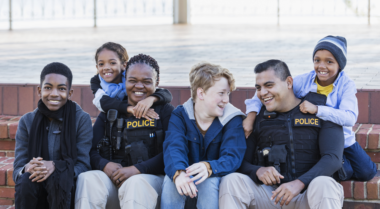 Community policing - two police officers hanging out with a group of four multi-ethnic children, sitting side by side on steps outside a building. The officers are an African-American woman in her 40s and an Hispanic man in his 20s. The children range in age from a 7 year old girl to a 14 year old teenage boy.