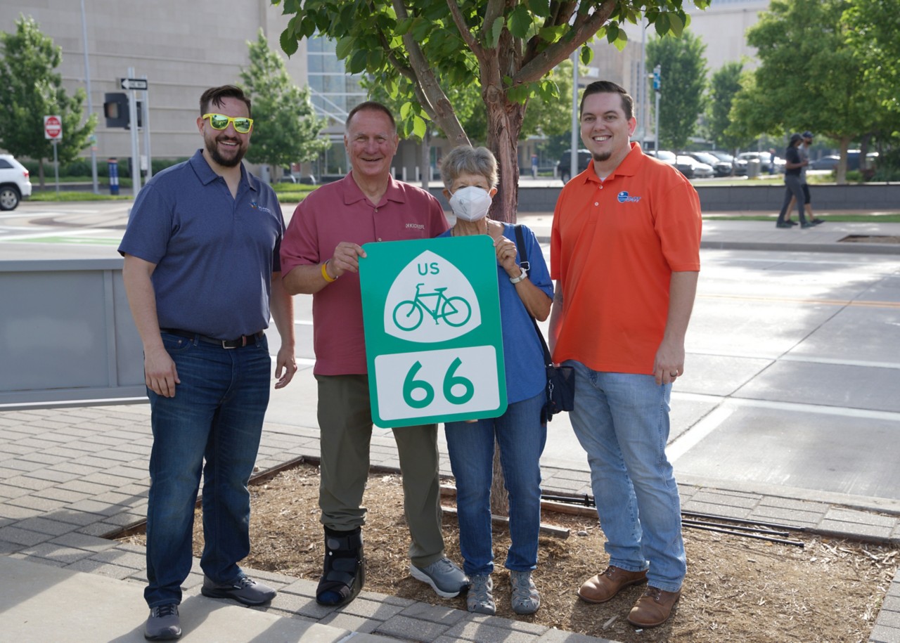 Four people standing outside and holding a road sign for U.S. Bicycle Route 66
