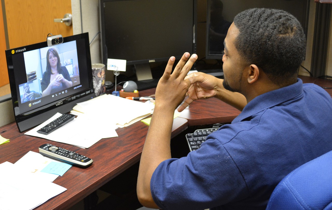 A man signing at his desk to a woman on a video call. 