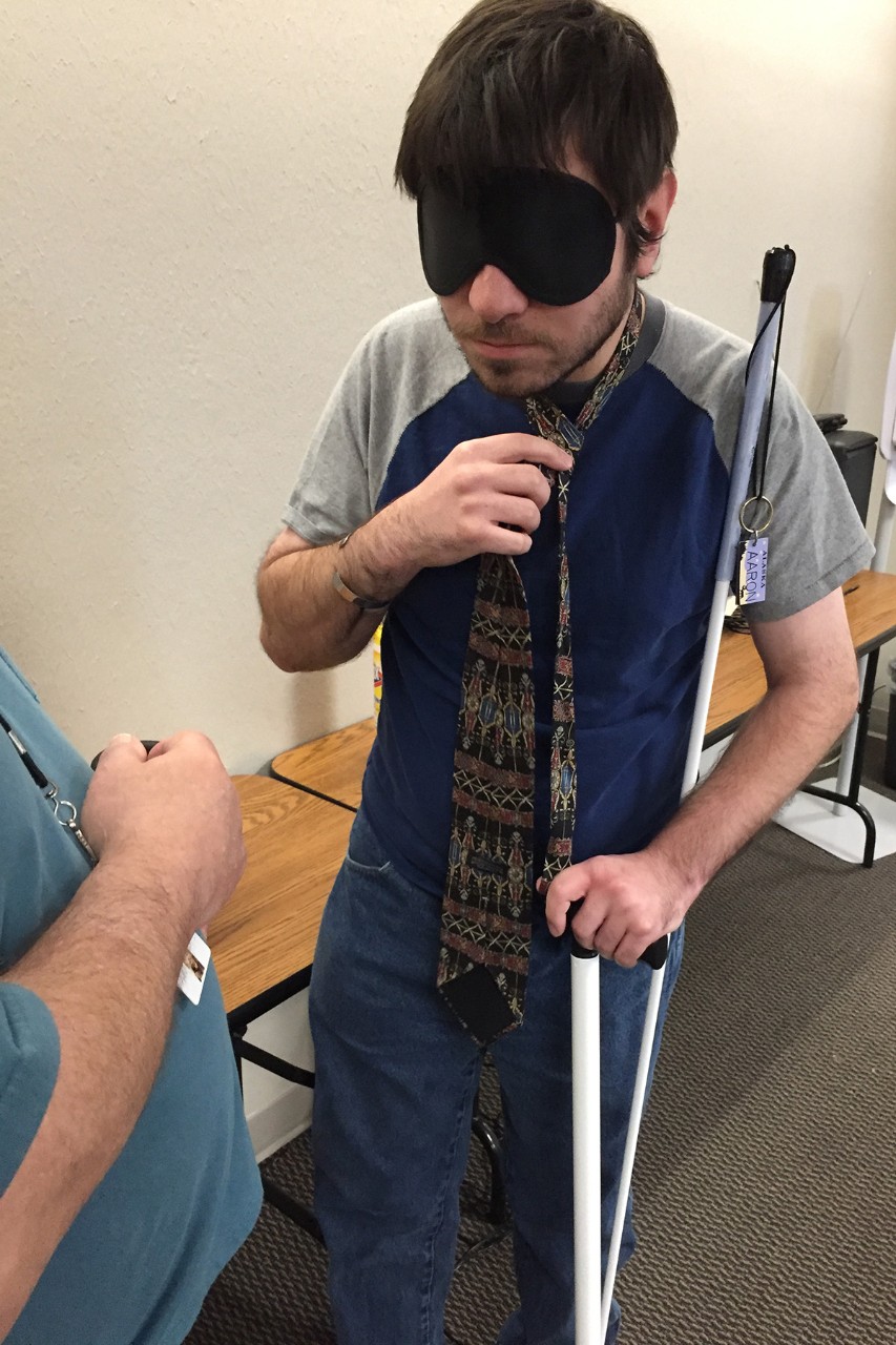 Blindfolded man holding white cane ties his tie.