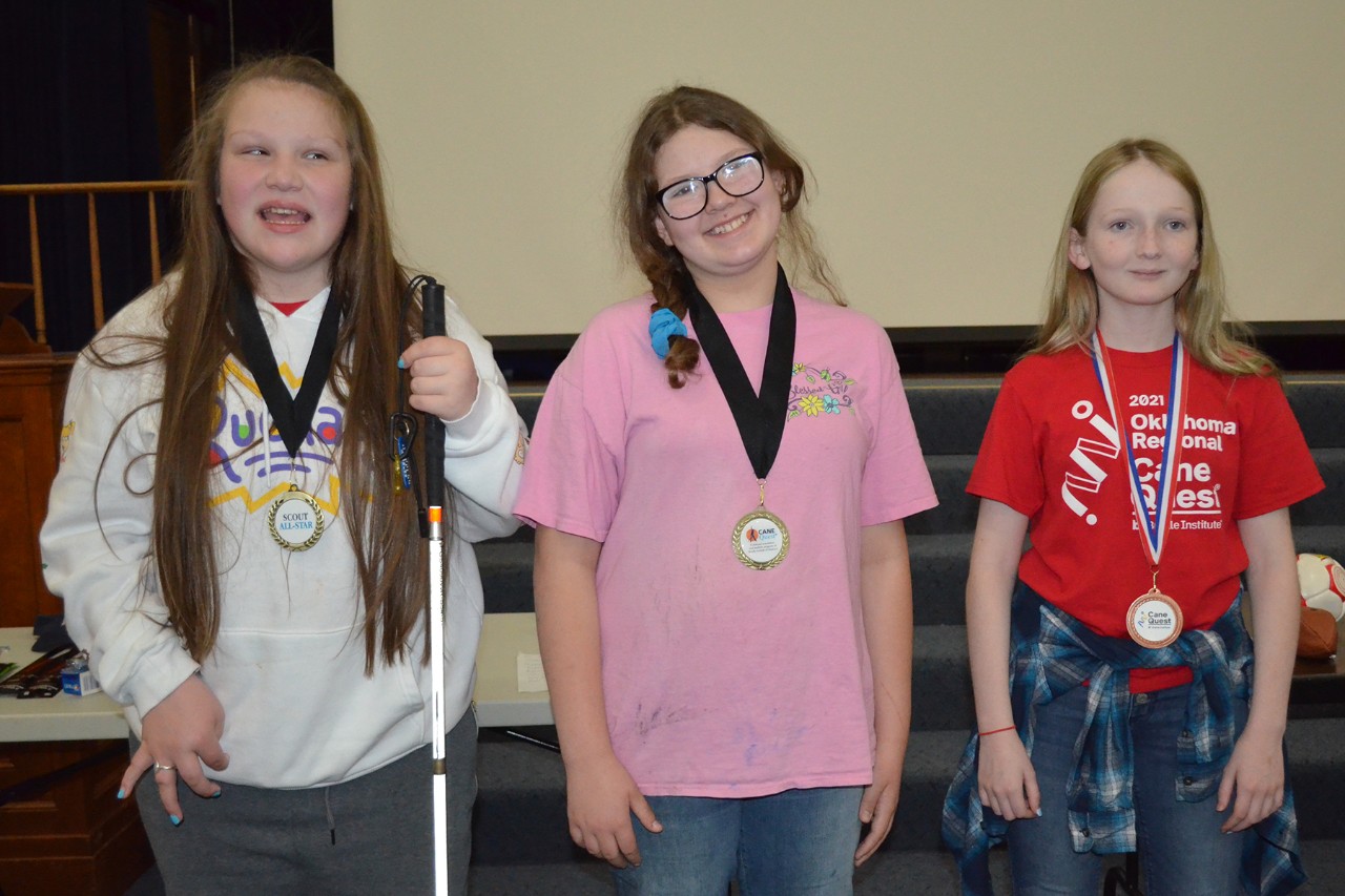 3 female students wear medals. 1 holds a long white cane.