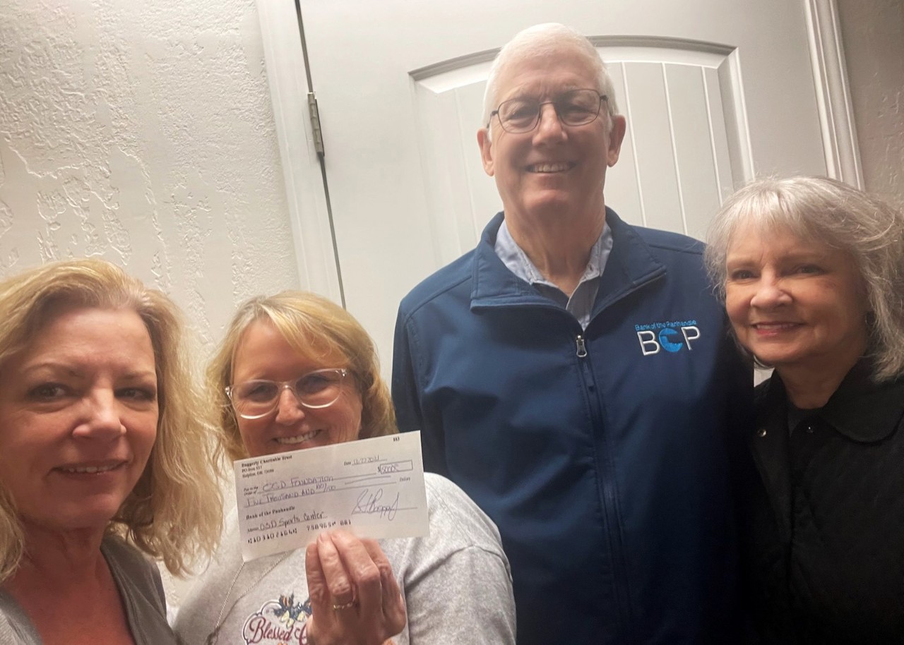 3 smiling women and 1 smiling man. Second woman holds up a check.
