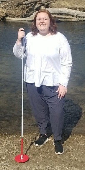 Woman holds white cane with red tip beside river.