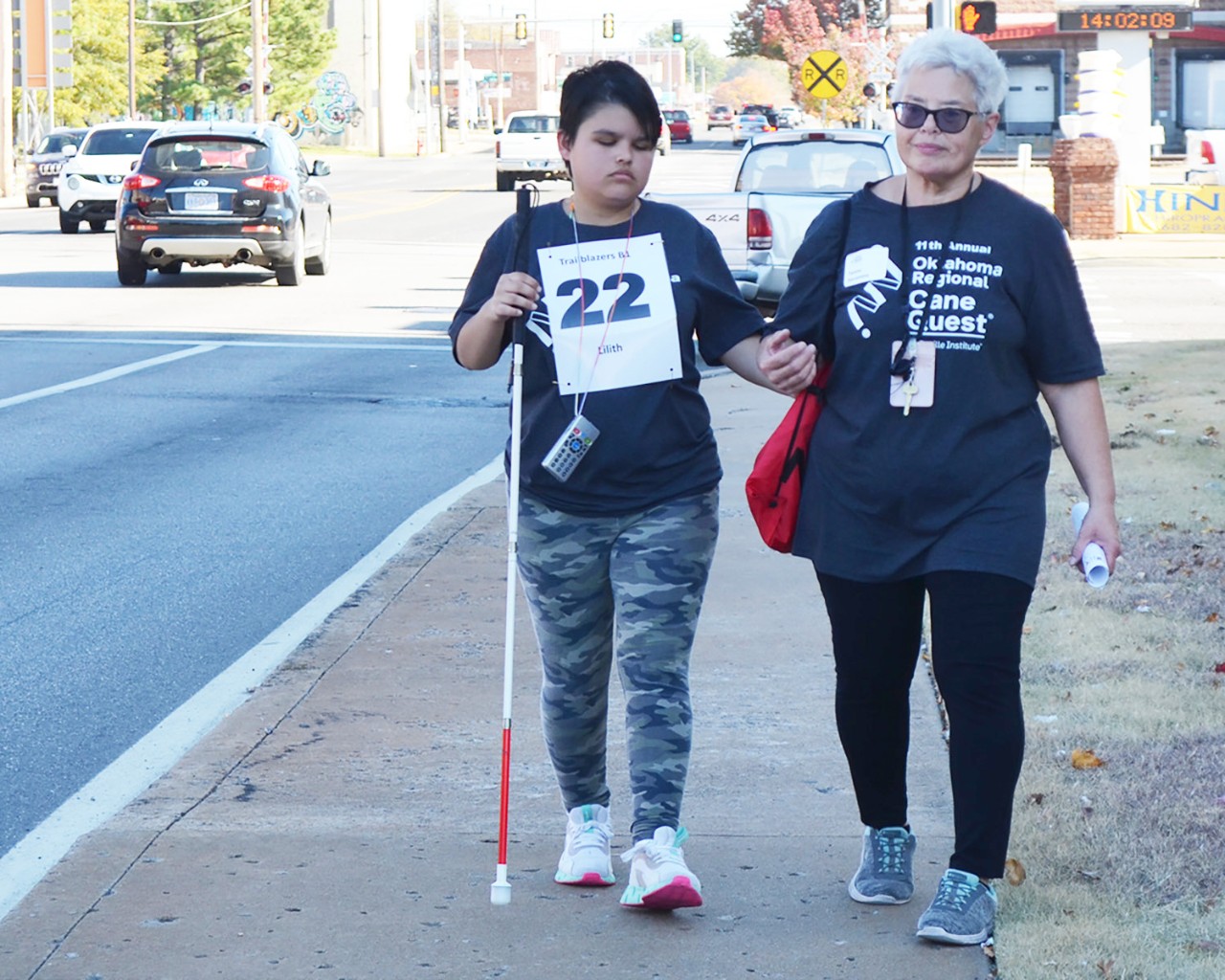 Young woman with white cane and 22 sign on chest walks with woman.