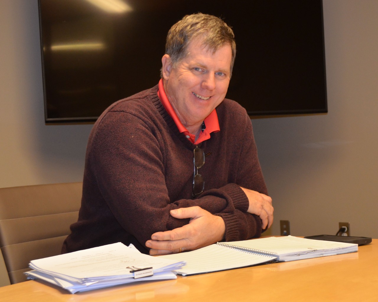 Smiling man seated a desk with arms folded on stacks of paper. 