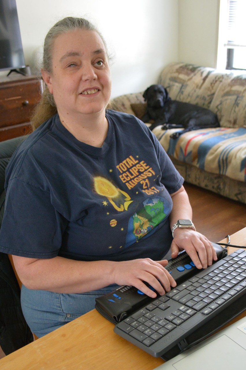 Smiling woman with hands on keyboard on desk with dog on couch in the background