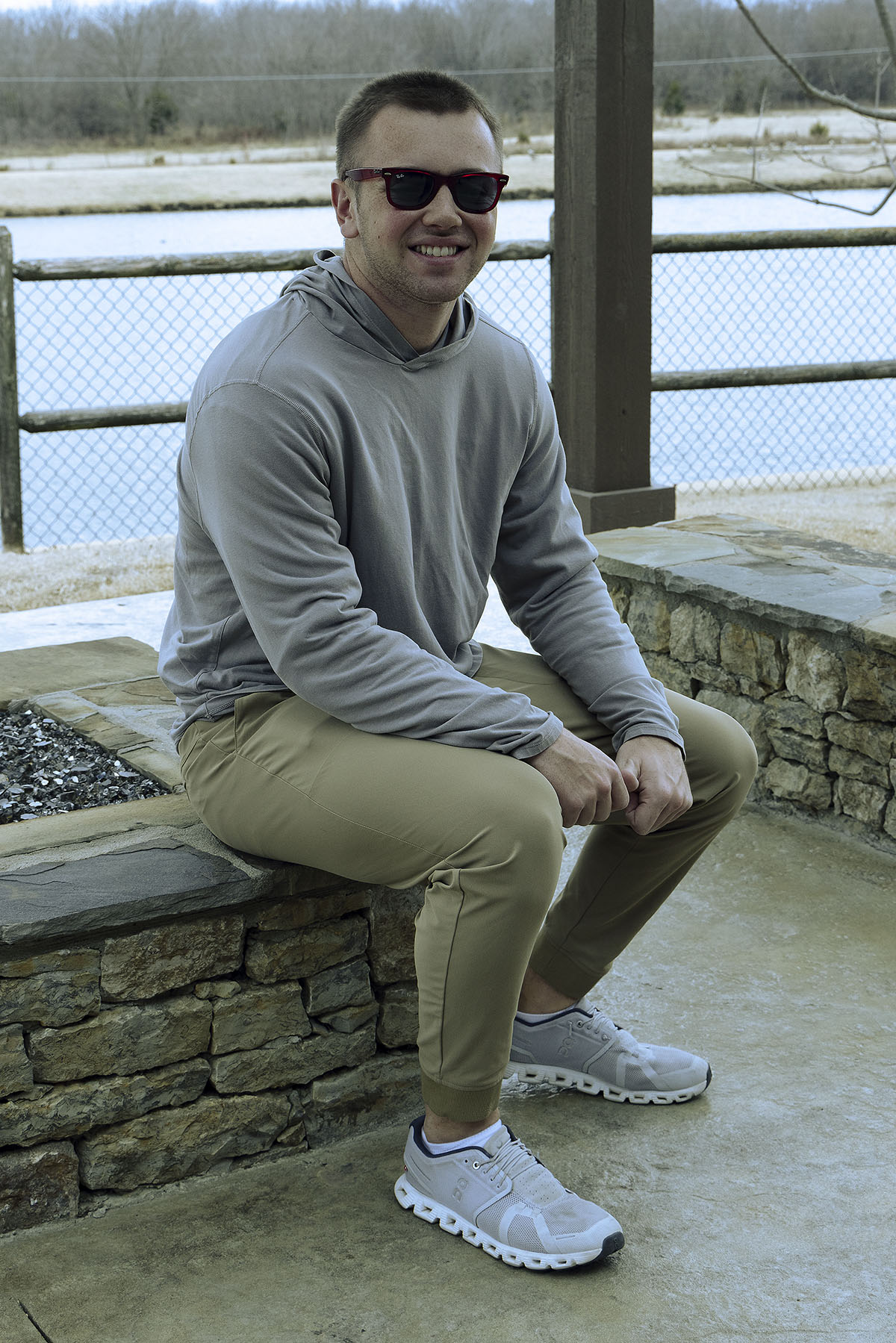 Smiling man wearing sunglasses sits on a stone wall in front of a lake.
