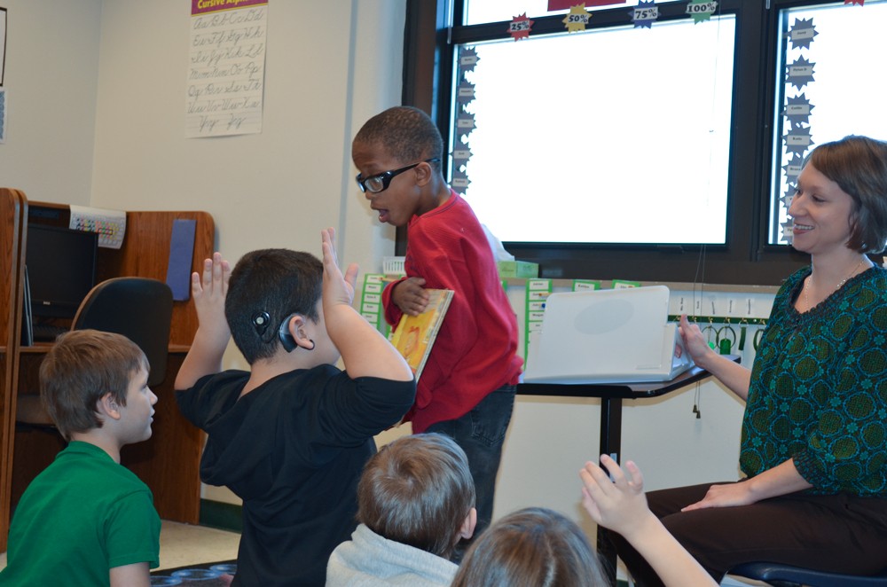 a little boy holds up a book for the class to see as he leads a read-a-long and the teacher smiles