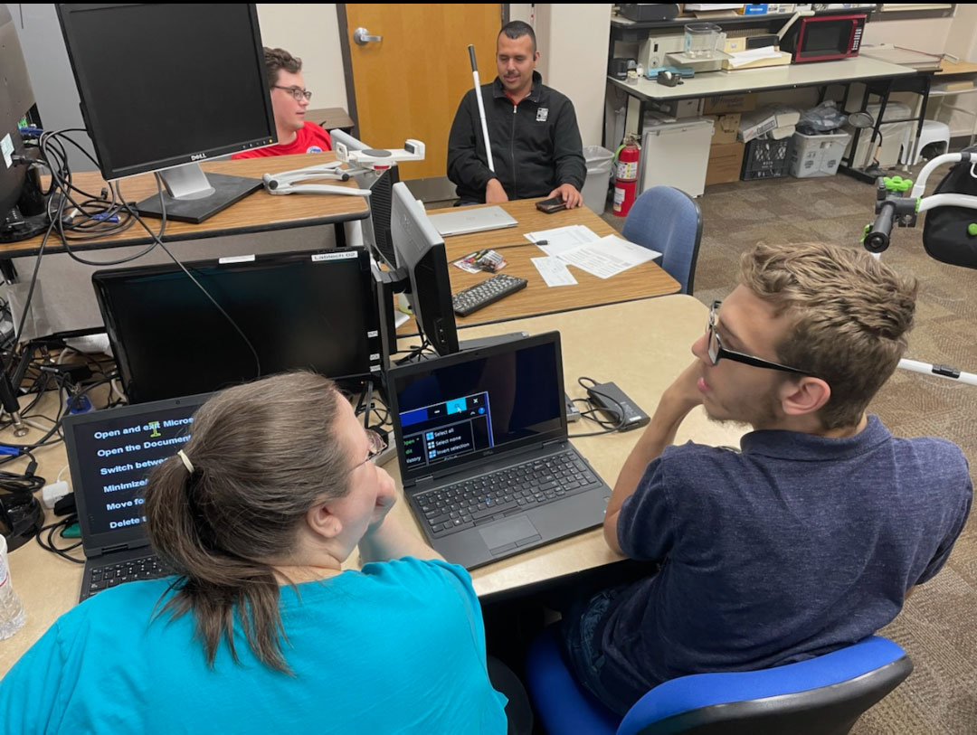 A group of four sitting a computer tables. One adult is talking with a VIBE student. Two students on the opposite side talk to each other. 
