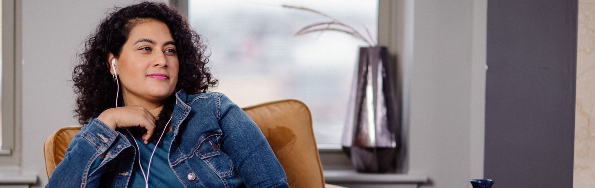 A woman relaxes in a chair. She holds a cell phone loosely in her left hand, which rests in her lap. Corded earbuds connected to the phone are in her ears and she looks off into the mid-distance.