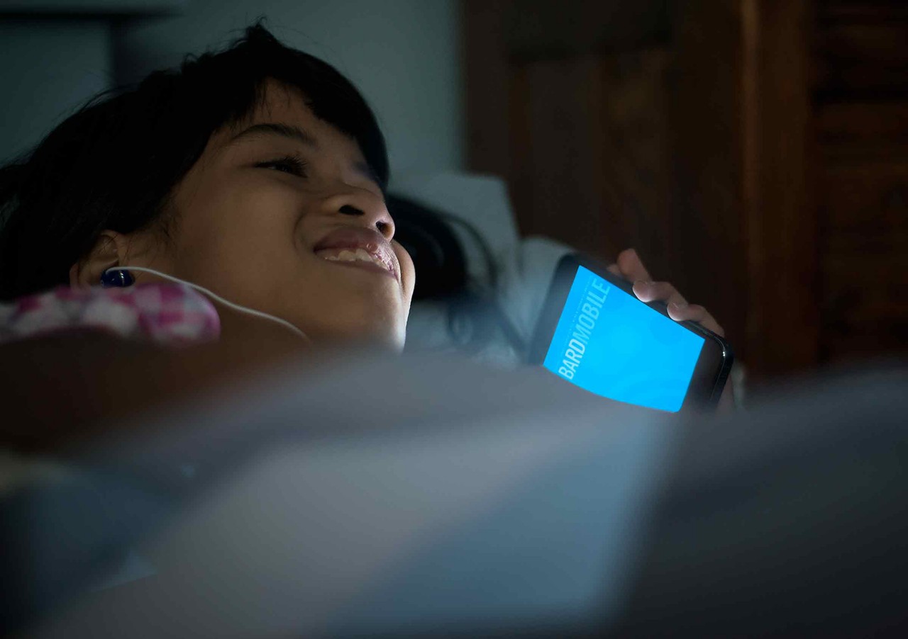 A middle-school aged girl holding a mobile phone lies reclined with corded earbuds in her ears. The phone screen displays the text "BARD Mobile" and shines a blueish light over the girl. The girl smiles softly. 