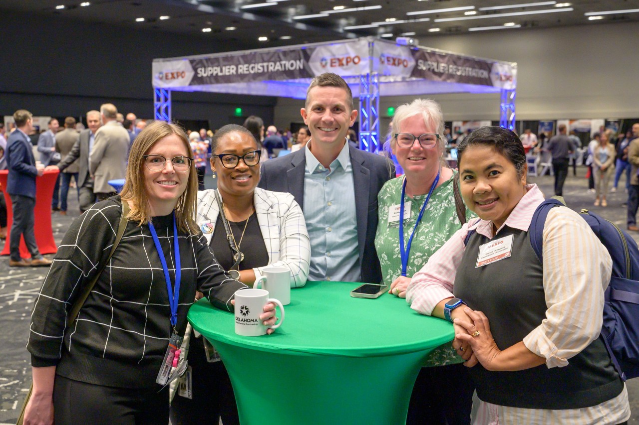 Five Education staff smiling around a ballroom bistro table in front of the Supplier Registration booth.