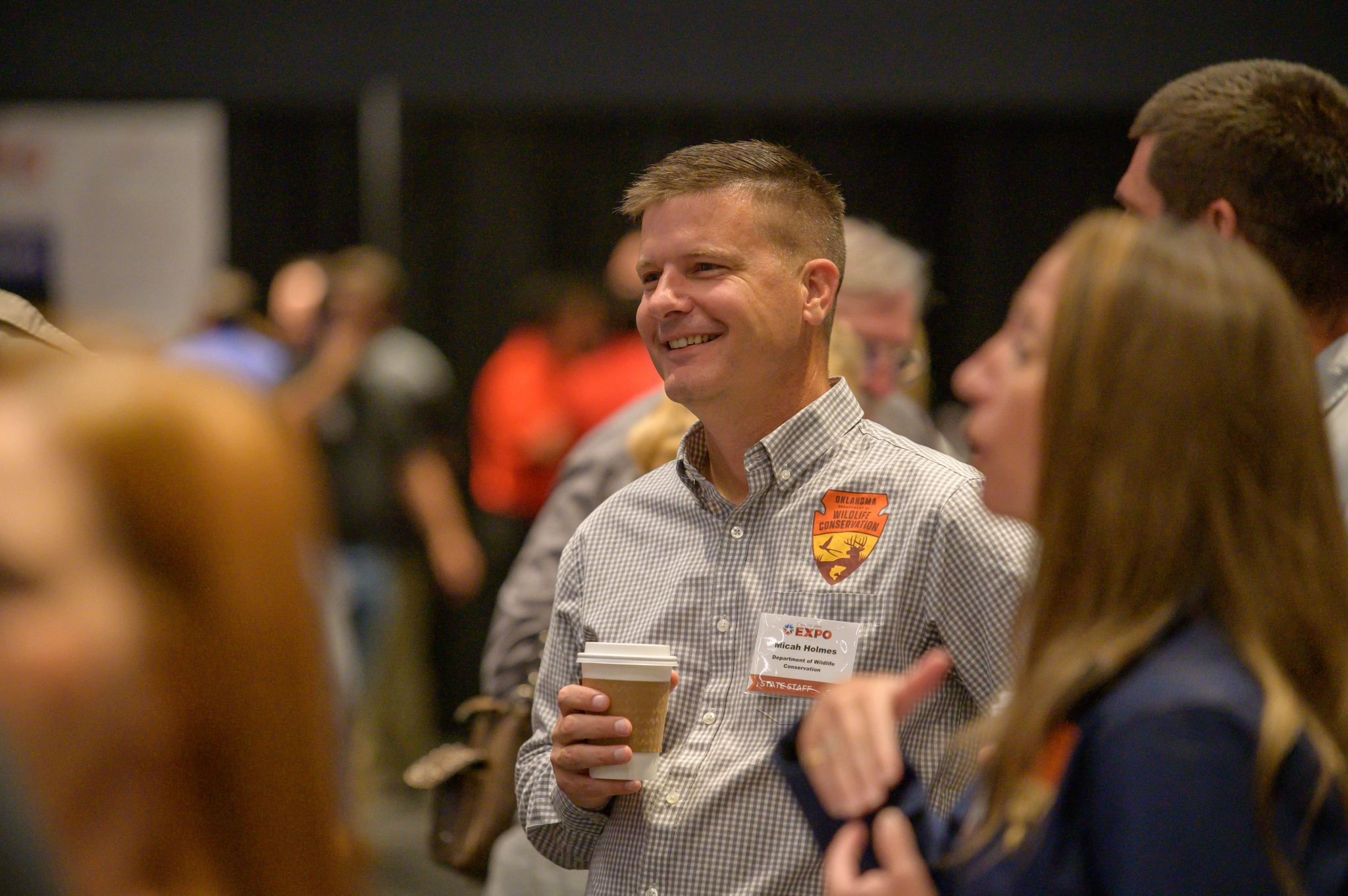 Micah Holmes smiles and holds coffee among other staff and attendees.