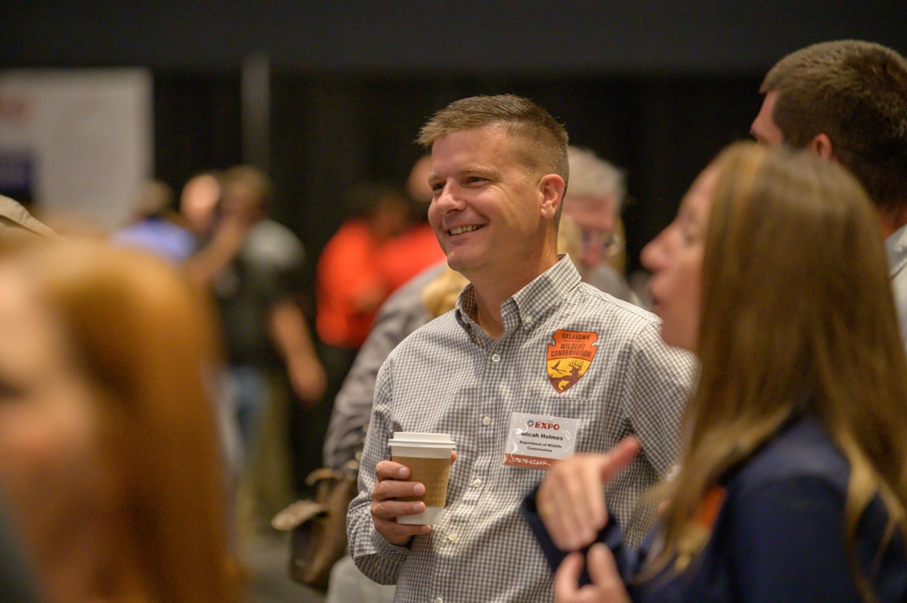 Micah Holmes smiles and holds coffee among other staff and attendees.