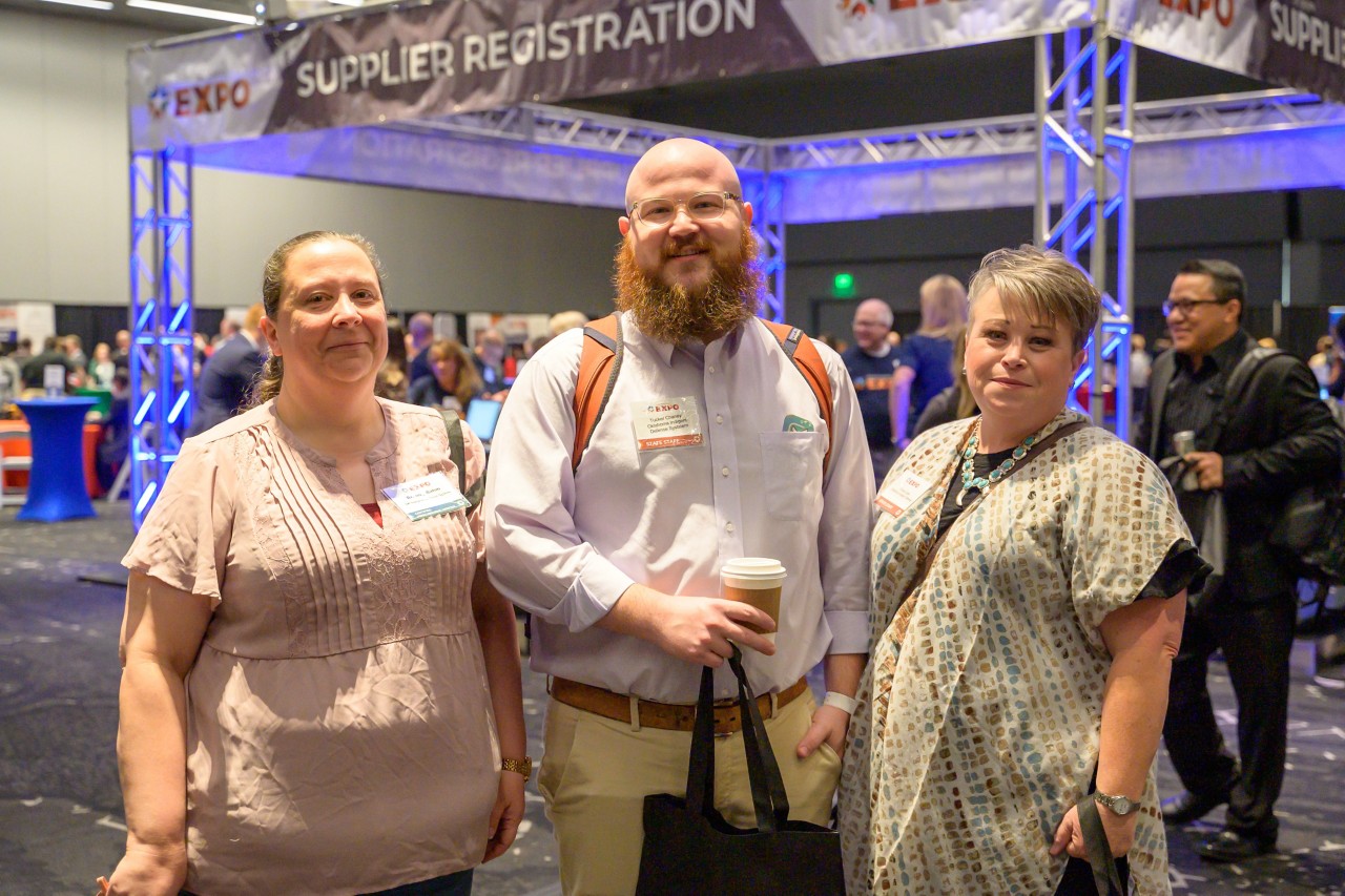 Three people stand and smile in the ballroom in front of the Supplier Registration booth.