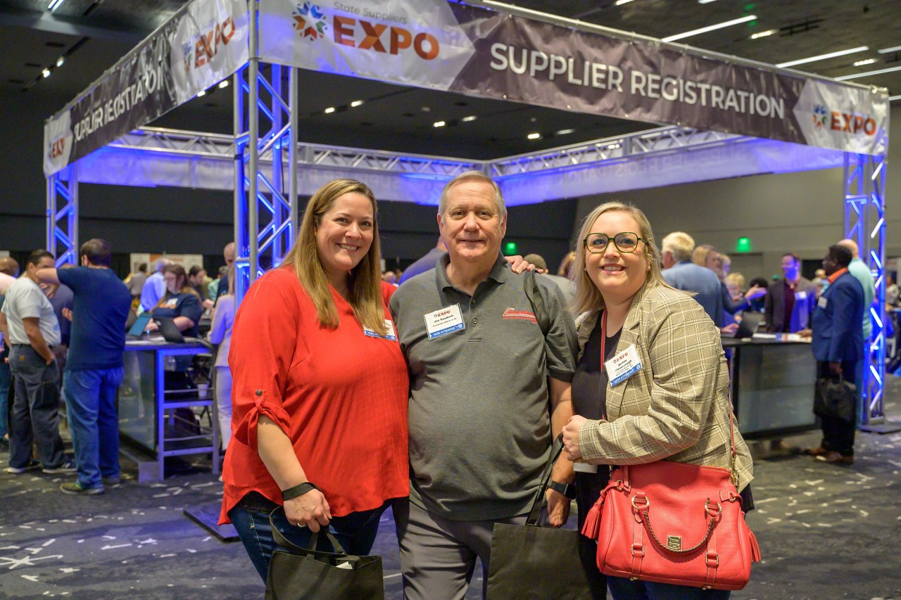 Three attendees smile and stand in front of the Supplier Registration booth.