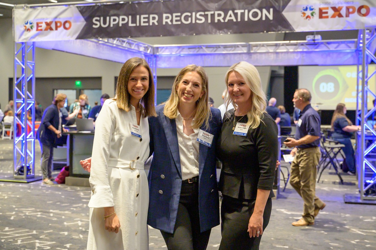 Three Thentia attendees smile in front of the Supplier Registration booth.